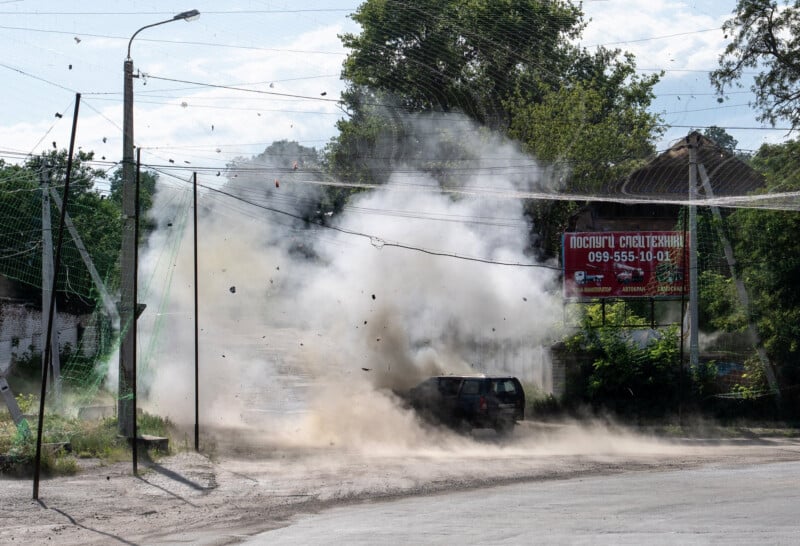 A car is engulfed in smoke and debris after an explosion on a city street, with dust and fragments in the air. Trees and a billboard are visible in the background.