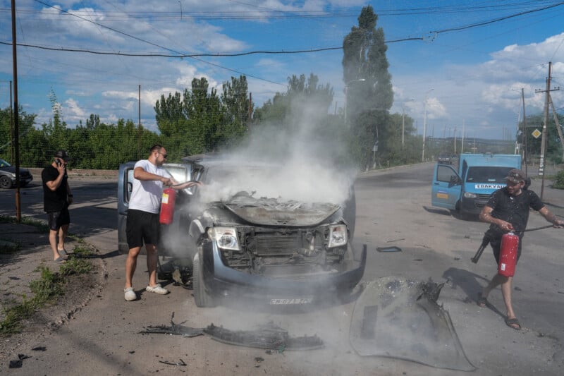 Three people use fire extinguishers to put out smoke coming from the engine of a damaged vehicle on a street, with debris scattered around and trees in the background.