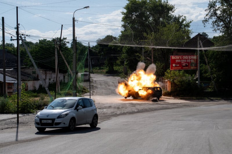 A burning car explodes on a deserted street as a white car drives by. Trees, power lines, and a red billboard are in the background on a sunny day.