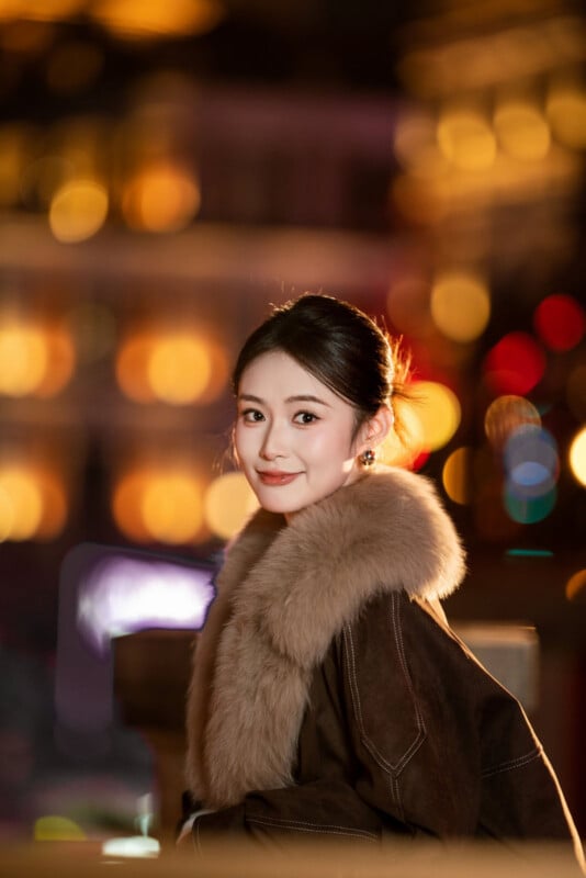 A young woman with dark hair in a bun, wearing a brown coat with a large fur collar, smiles at the camera. She is outdoors at night, with colorful blurred city lights in the background.