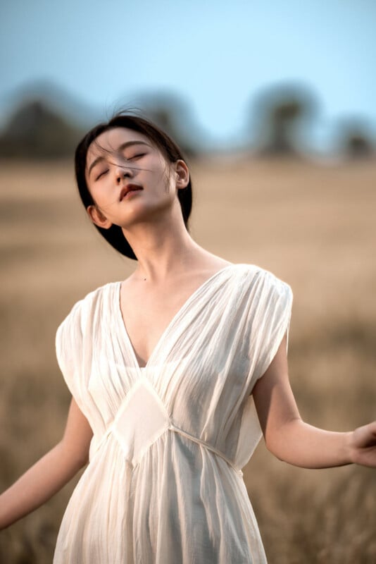 A young woman in a flowing white dress stands in a sunlit field with her eyes closed, head tilted back, and hair gently blowing in the breeze. The background is softly blurred.