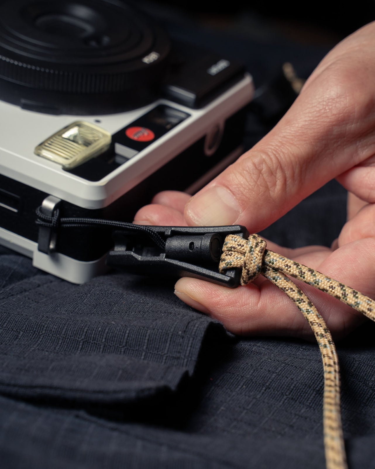 A close-up of a hand attaching a beige and black braided camera strap with a clasp to a silver and black camera resting on a dark fabric surface.
