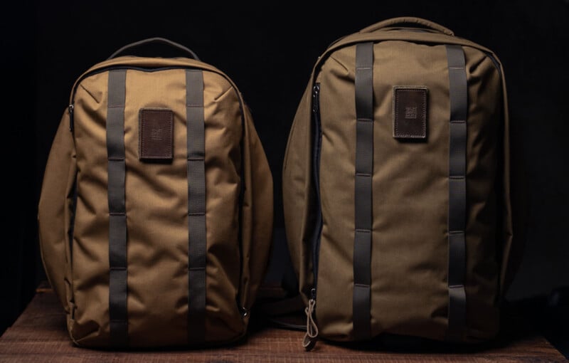 Two brown backpacks with black straps and square leather patches sit side by side on a wooden surface against a dark background.
