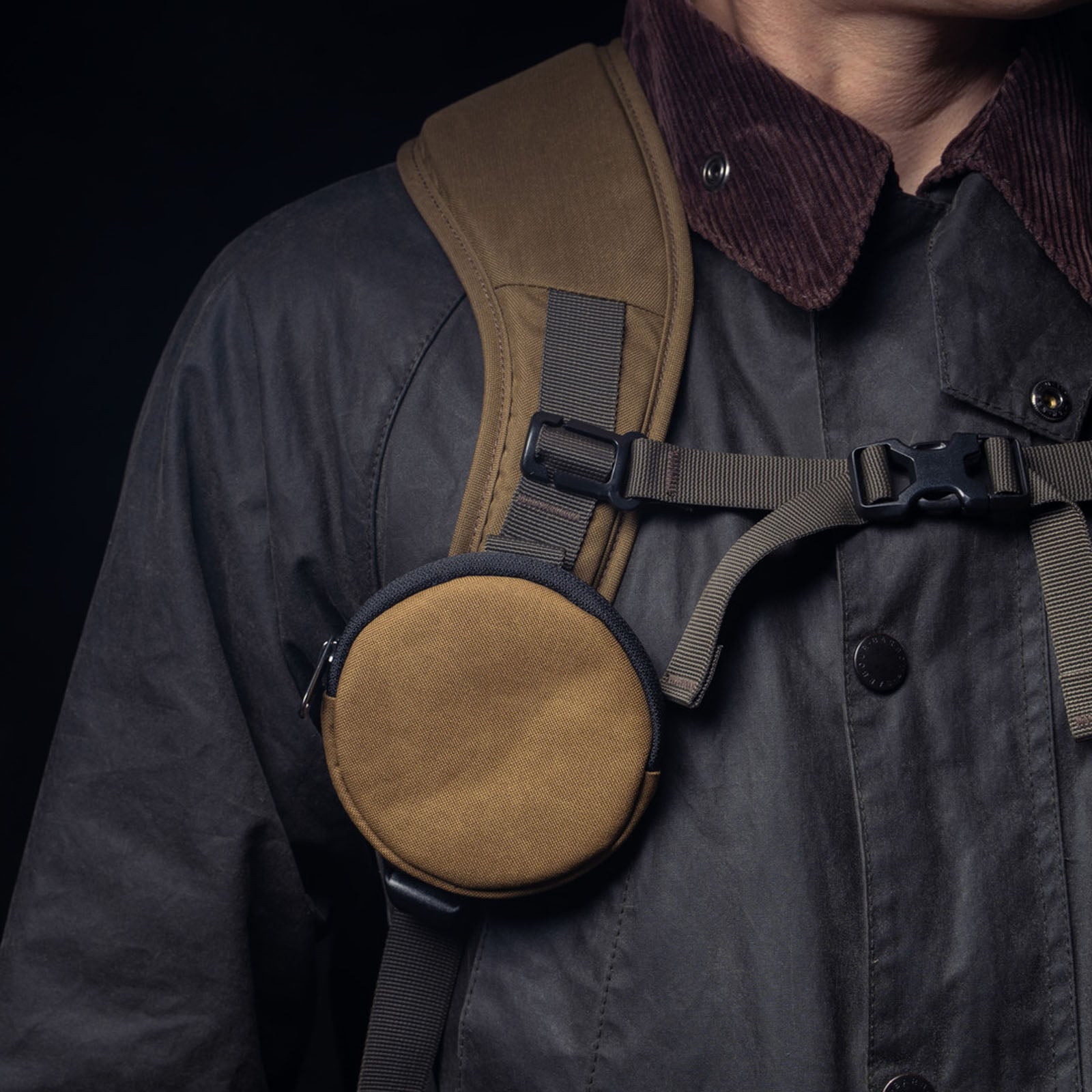 A person wearing a dark jacket and brown strap backpack with a round, tan pouch attached to the chest harness.