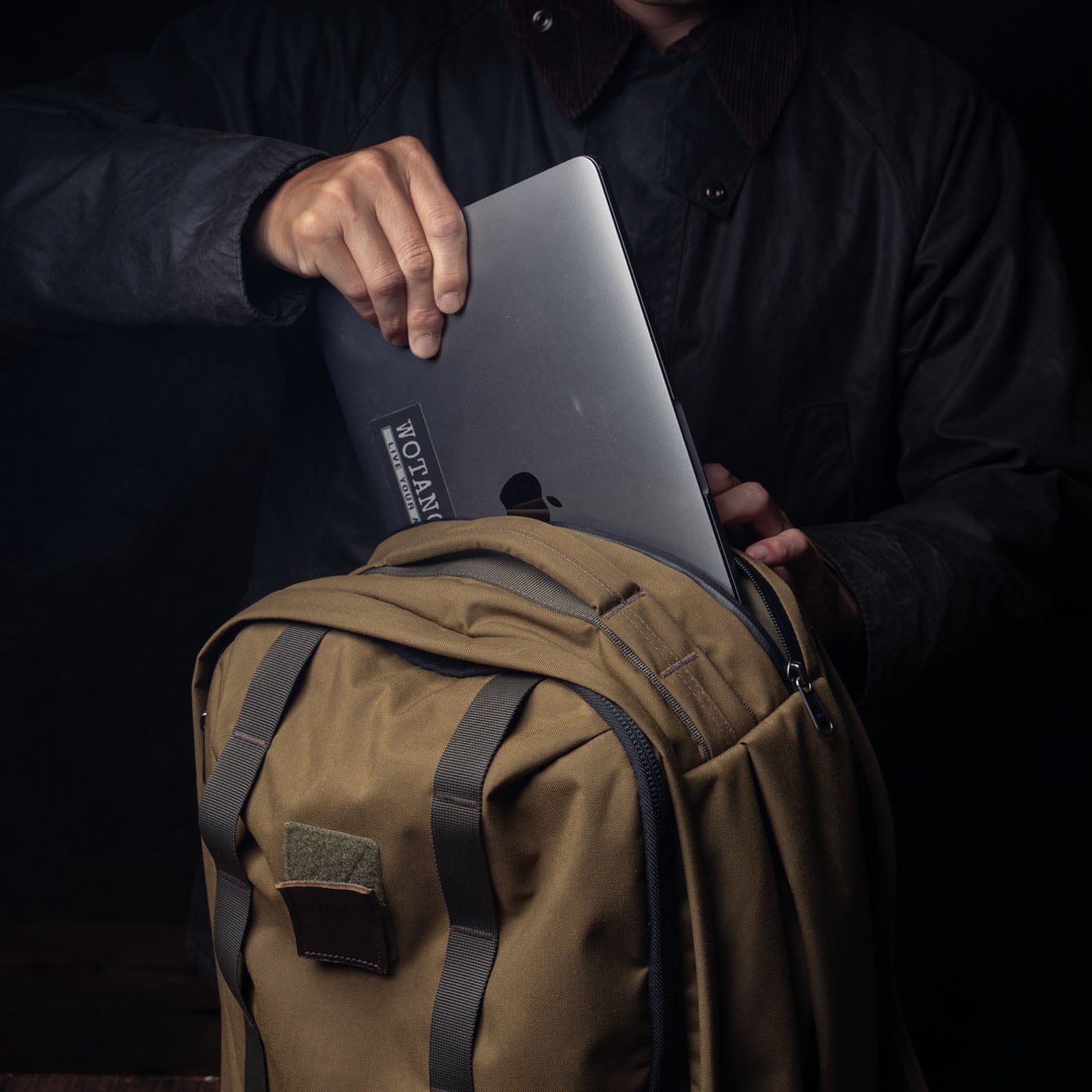 A person wearing a dark jacket places a silver laptop into a tan backpack. The laptop has a visible sticker. The scene is dimly lit, with a dark background.