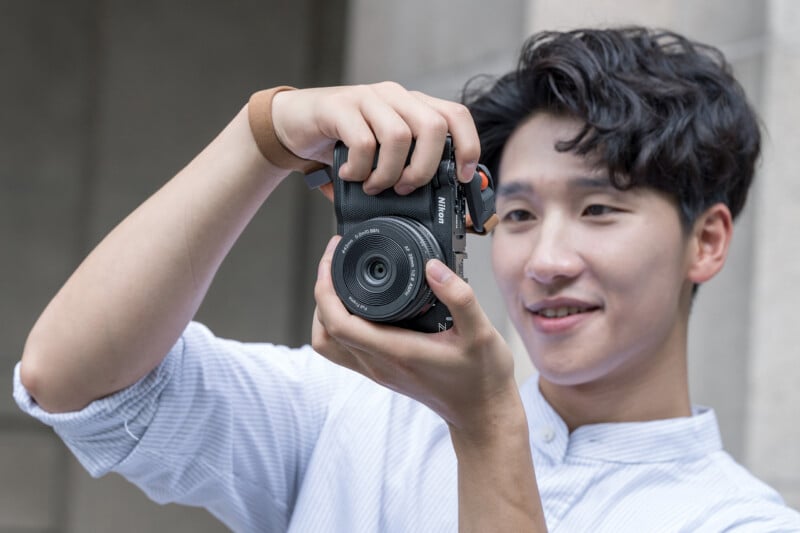 A young person in a light shirt smiles while holding a camera up to take a photo outdoors against a neutral stone background.