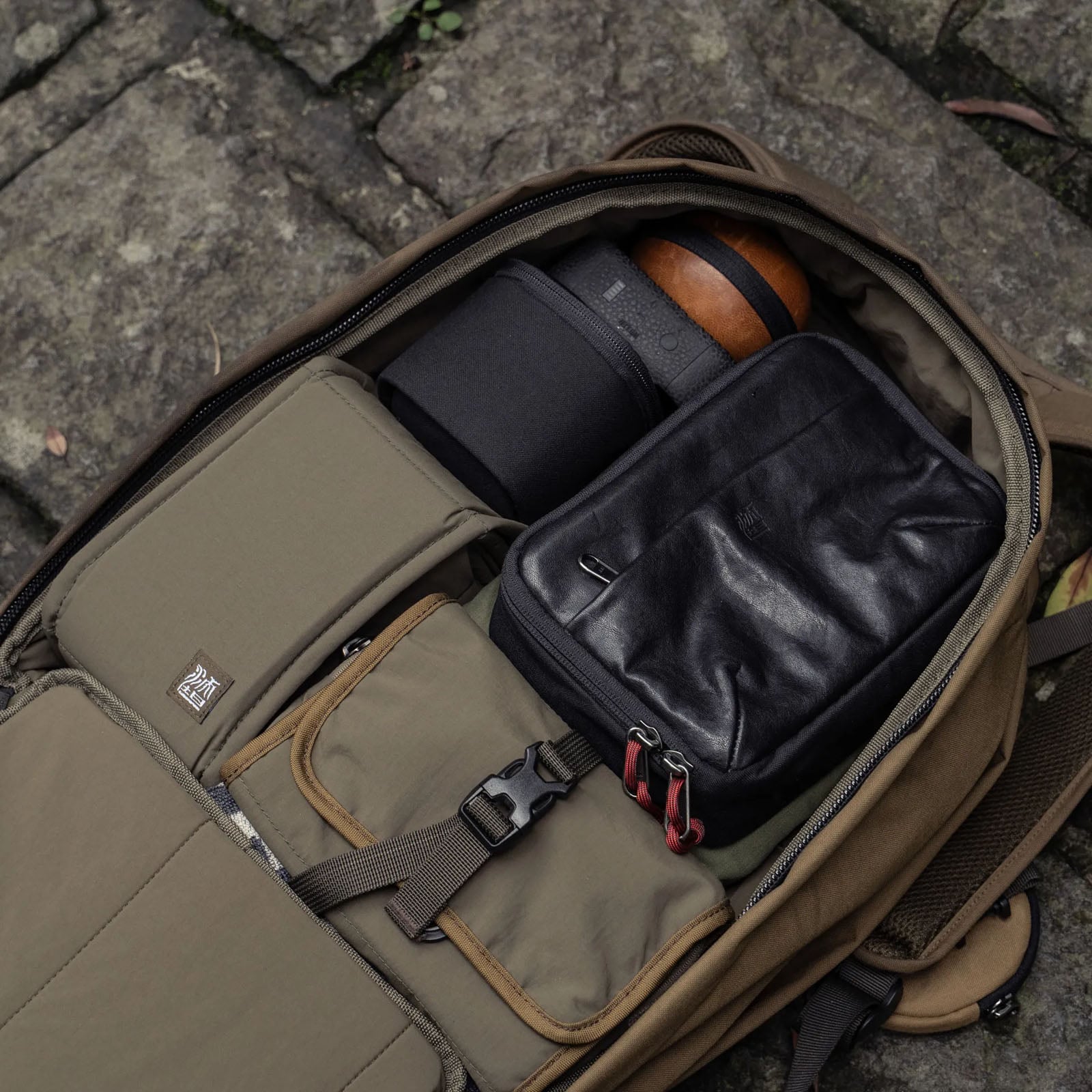 Open backpack on stone ground, containing a camera lens in a black case, a black leather pouch, and several organized compartments with beige and brown fabric pouches and straps visible.