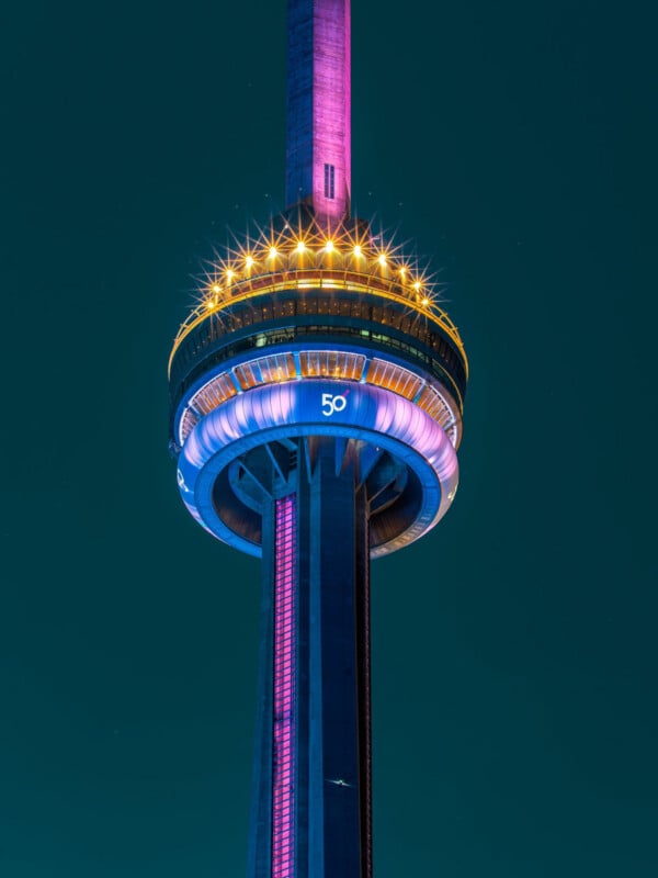Close-up view of the CN Tower at night, illuminated with vibrant pink, purple, and yellow lights. The number "50" is displayed in blue, marking the tower’s 50th anniversary. The sky is dark in the background.