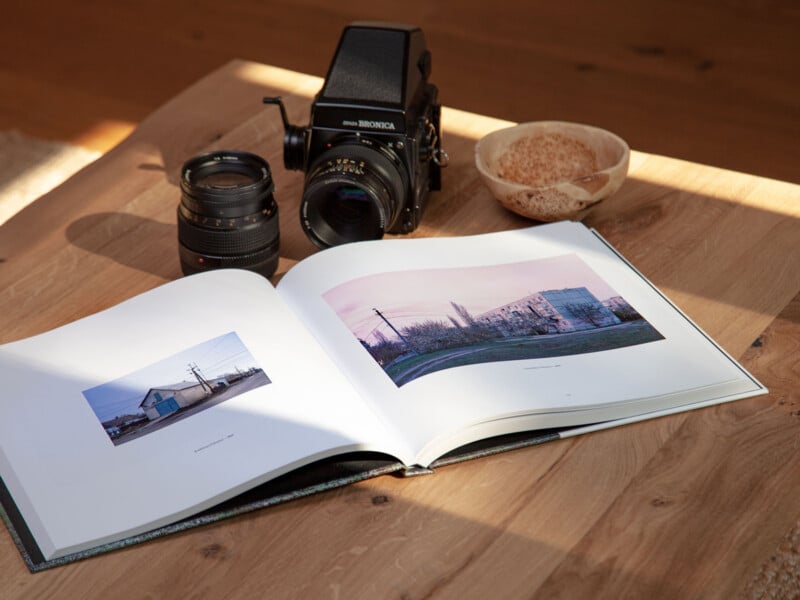 An open photo book displaying landscape images rests on a wooden table with a camera, extra lens, and a bowl of coffee or tea nearby, all illuminated by natural sunlight.