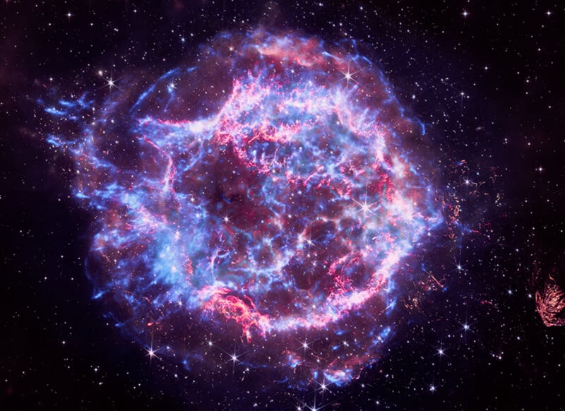 A colorful nebula in deep space glows with blue and pink clouds of gas and dust, surrounded by countless small white stars against a dark backdrop.