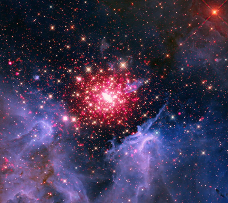 A vibrant star cluster shines in deep space, surrounded by glowing clouds of blue and purple gas, with numerous red and white stars scattered throughout the scene.