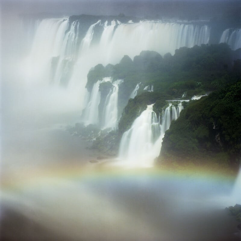 A misty view of large waterfalls cascading over lush green cliffs, with a faint rainbow visible through the mist near the base of the falls.