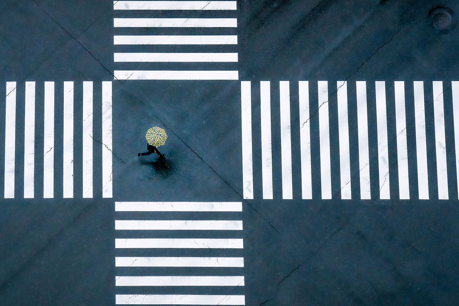 A person holding a yellow umbrella crosses the center of an empty intersection with bold white crosswalk lines, seen from above on a wet, dark road.