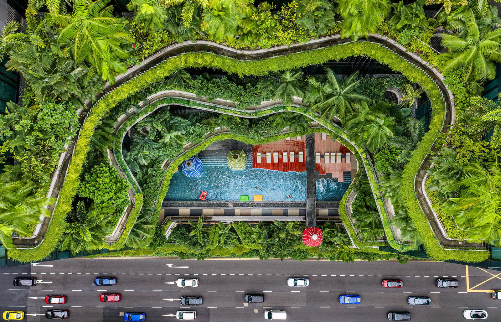 Aerial view of a lush rooftop garden with a swimming pool, palm trees, lounge chairs, and colorful umbrellas, situated above a busy multi-lane road with moving cars.