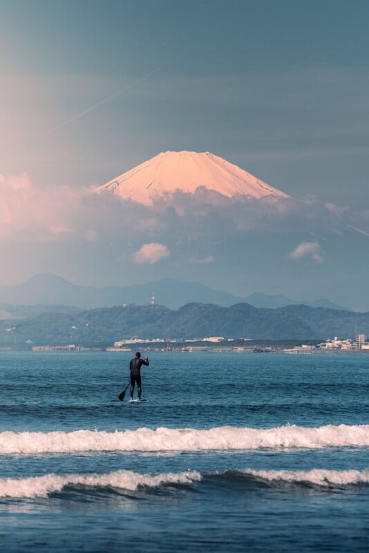 A person paddleboarding on calm water with gentle waves in the foreground, and Mount Fuji towering in the background under a partly cloudy sky.