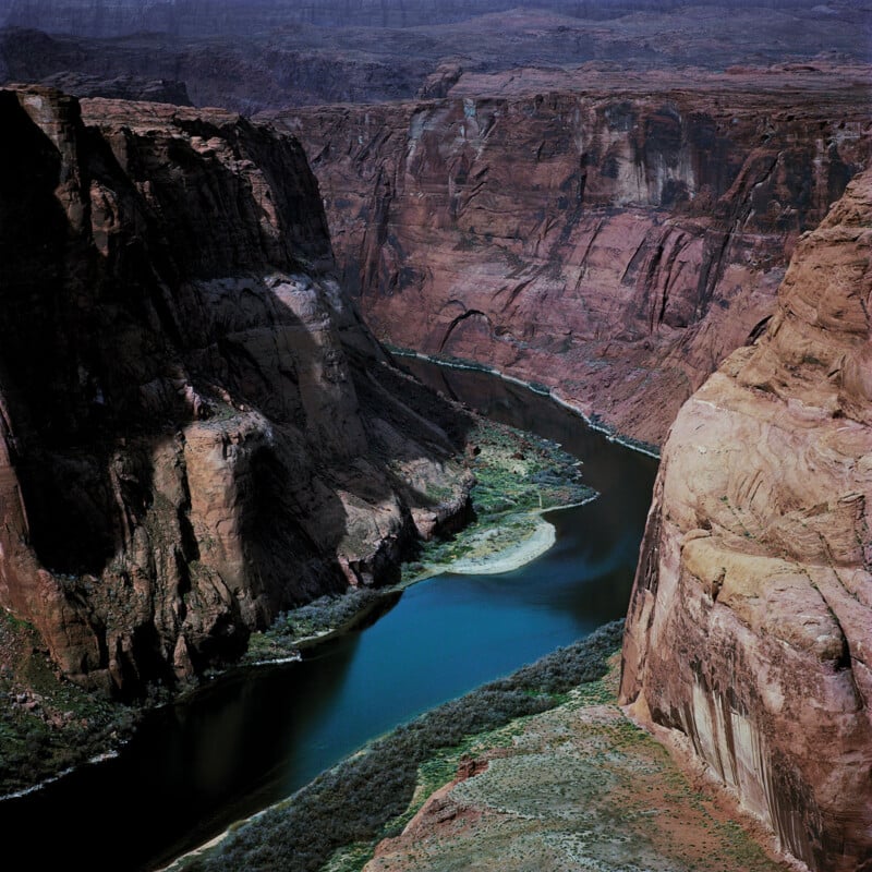 A winding river flows through a deep canyon with steep, rugged red rock cliffs under a hazy sky, with sparse vegetation lining the water’s edge.