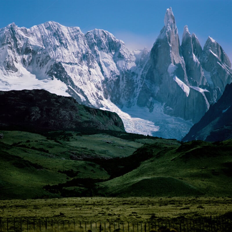 Snow-covered jagged mountains rise behind green rolling hills and a fence under a blue sky.