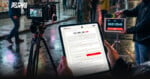 A person holds a tablet displaying a film production form on a rainy street set, with a camera on a tripod and a clapperboard in the background. 