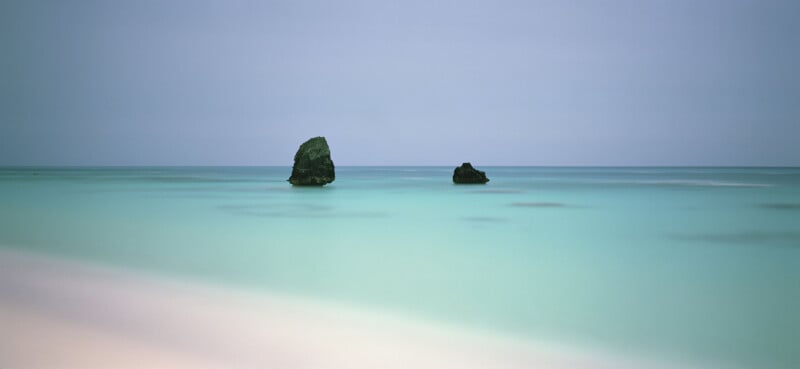 Two dark rocks emerge from calm, turquoise water under a pale blue sky, with soft, sandy beach in the foreground. The scene is minimalist and tranquil.