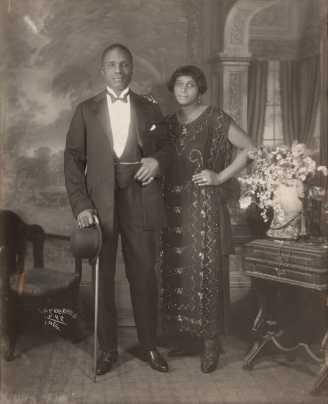 A 1924 studio portrait by James Van Der Zee of a Black couple standing together, the man in a tuxedo holding a top hat and cane, and the woman in a beaded flapper-style dress.