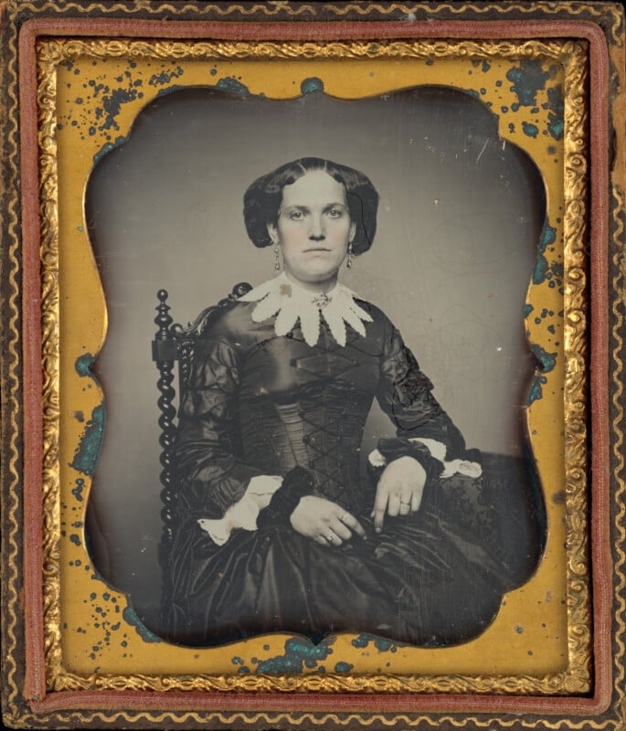 A daguerreotype portrait of a woman in a dark, long-sleeved dress with a white lace collar, seated in a carved wooden chair.