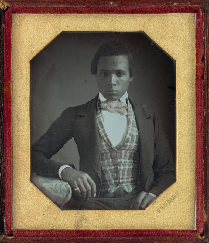 A daguerreotype portrait of a young Black man wearing a dark coat, a plaid vest, and a light-colored bow tie, set in a gold-toned octagonal frame with red velvet lining.
