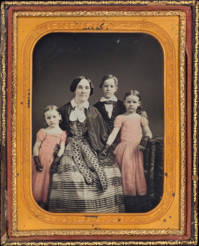 A studio portrait of a woman seated with a young boy and two young girls, all wearing formal mid-19th-century attire, set within an ornate gold-colored frame.