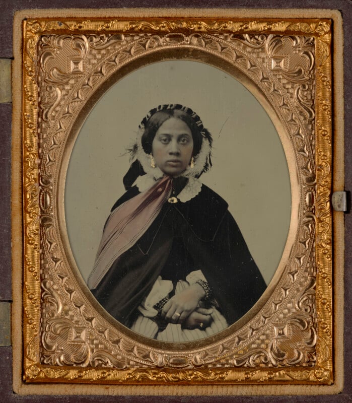A hand-tinted daguerreotype portrait of a woman wearing a dark cape, a white lace-trimmed bonnet, and jewelry, set within an ornate, rectangular gold-colored frame.