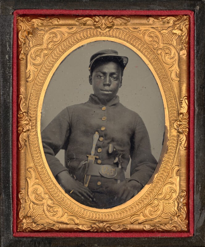 A tintype portrait of a young Black soldier in a Union uniform, wearing a forage cap and a belt buckle with the letters "US," framed in an ornate gold oval.