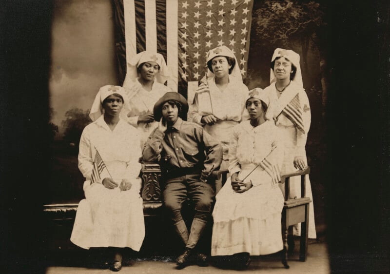A group of six Black women posing in front of an American flag, with five dressed as Red Cross nurses and one seated in the center wearing a military-style uniform.