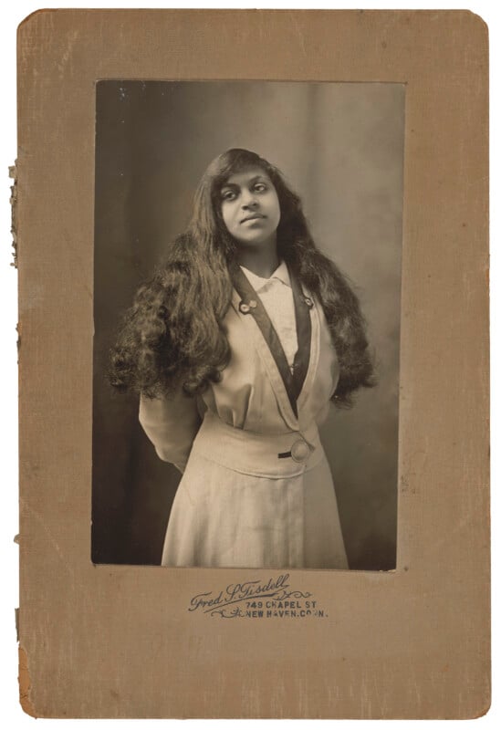 A vintage portrait of a young woman with long, dark, wavy hair, wearing a light-colored jacket with a dark lapel, mounted on a card printed with "Fred S. Tisdell, 749 Chapel St., New Haven, Conn.