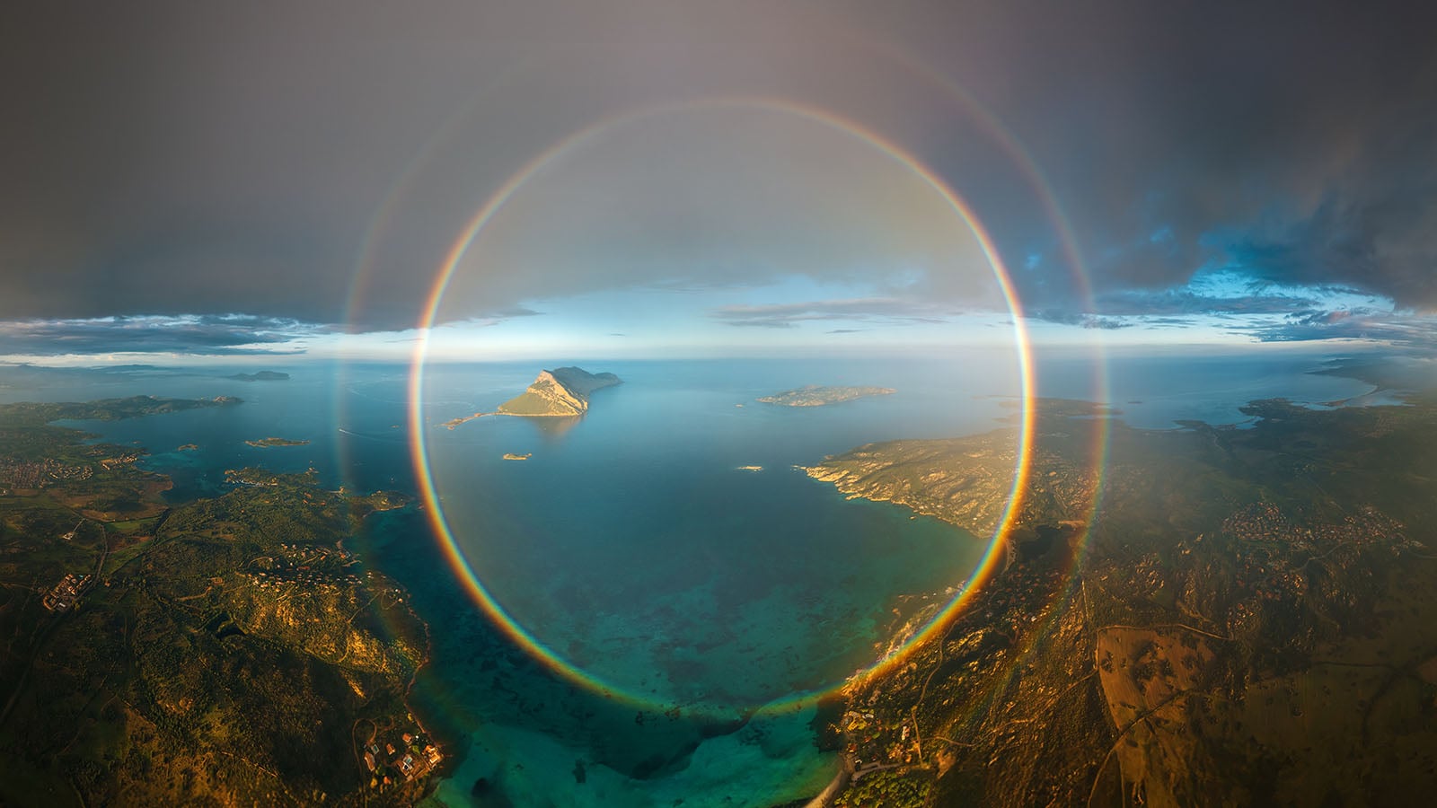 A stunning aerial view of a coastal landscape with a large, circular rainbow (glory) surrounding an island in the distance, under dramatic skies with clouds and blue water below.
