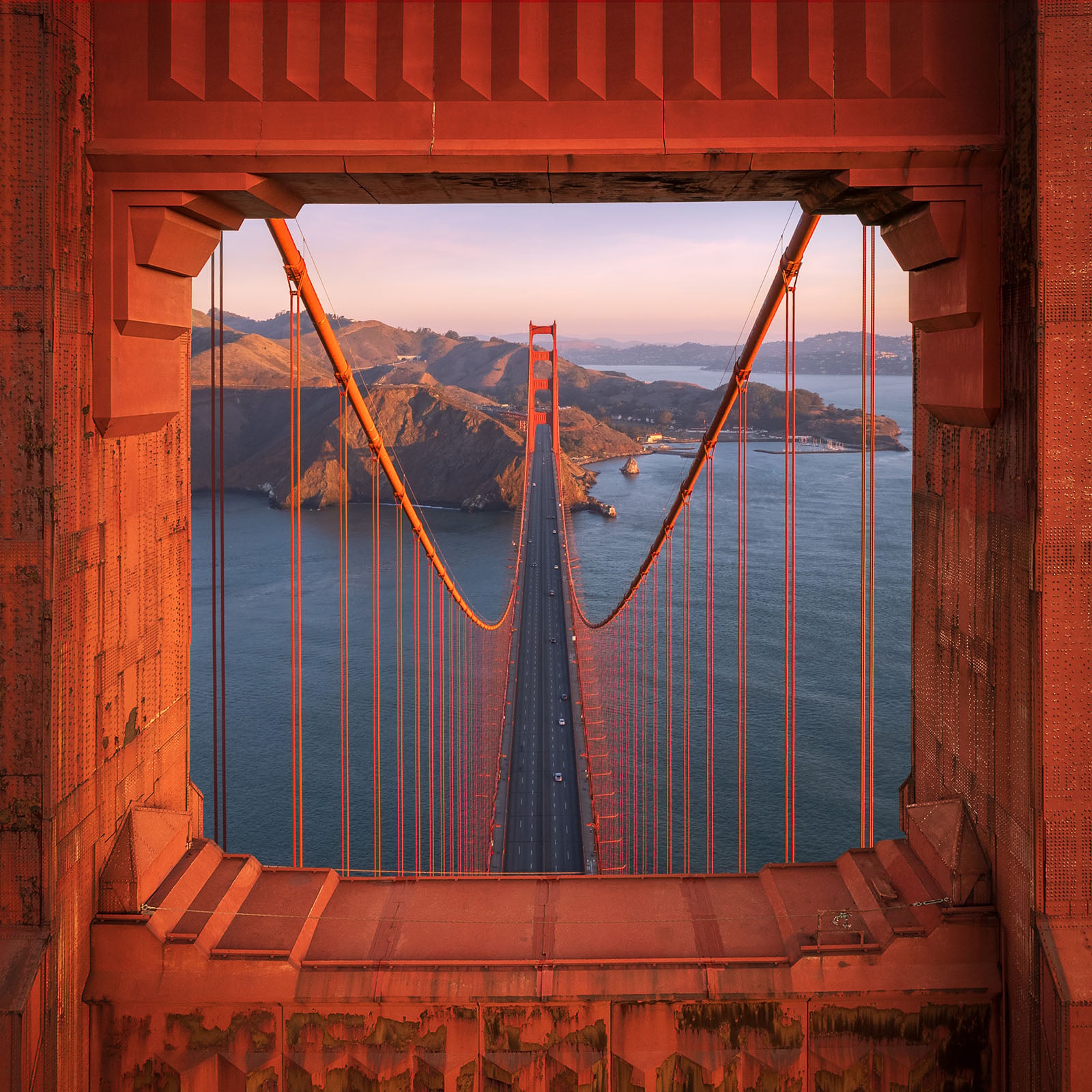View of the Golden Gate Bridge at sunset, framed by a rust-colored rectangular window, with the ocean and hills in the background.