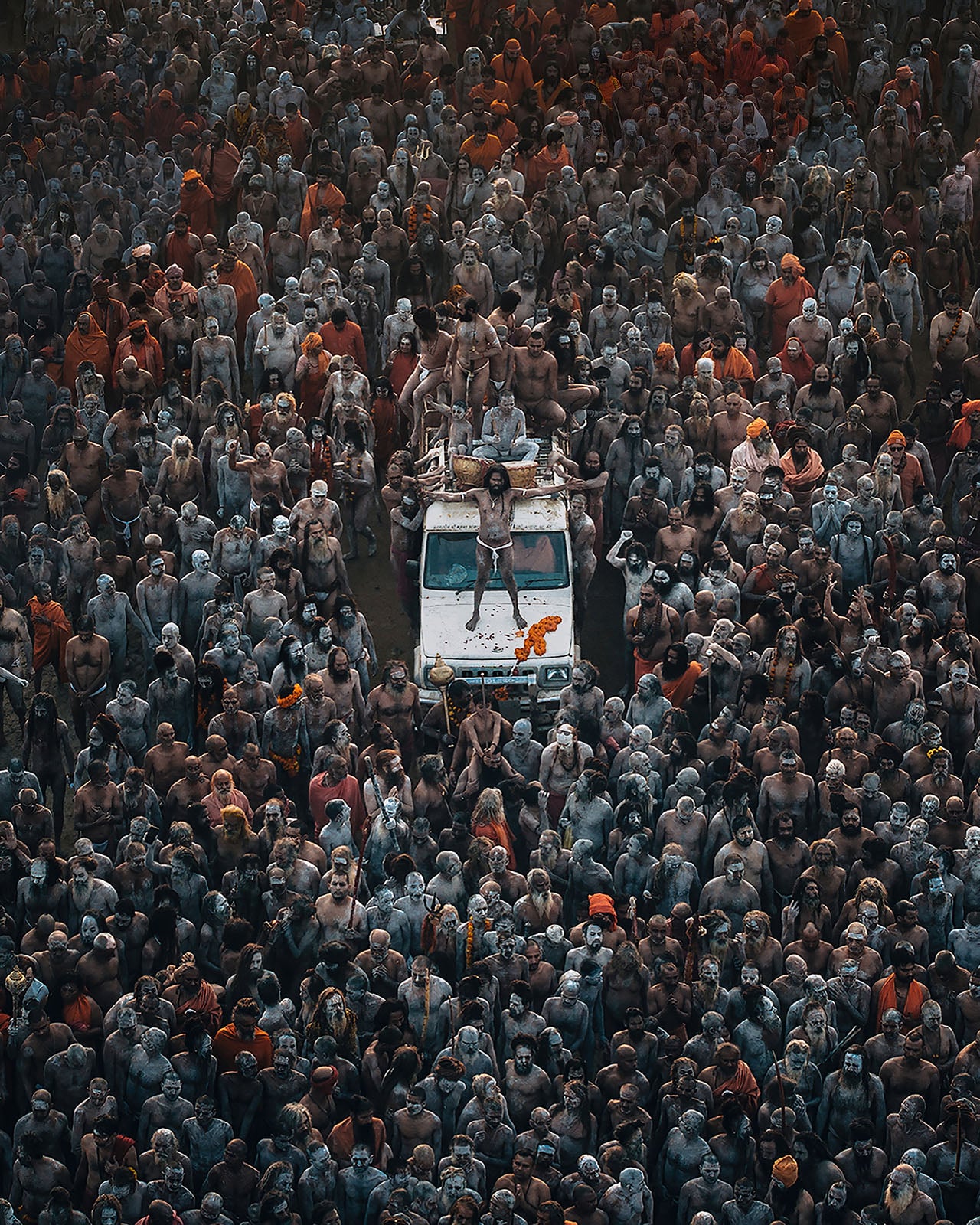 A large crowd of people, many with bodies painted in ash or white, gather around and on top of a white vehicle during a cultural or religious event. One person stands on the vehicle, raising their arms.