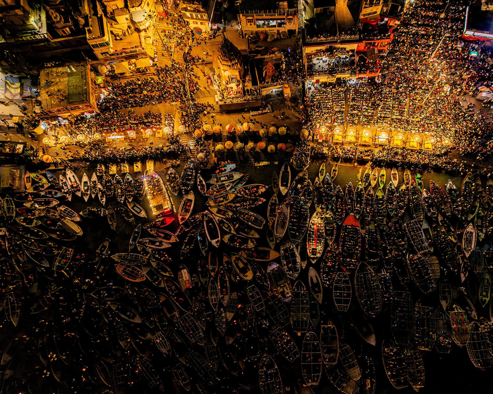 Aerial view of a crowded riverside at night, with dozens of boats packed closely together in the water and a large gathering of people illuminated by warm lights along the shore.