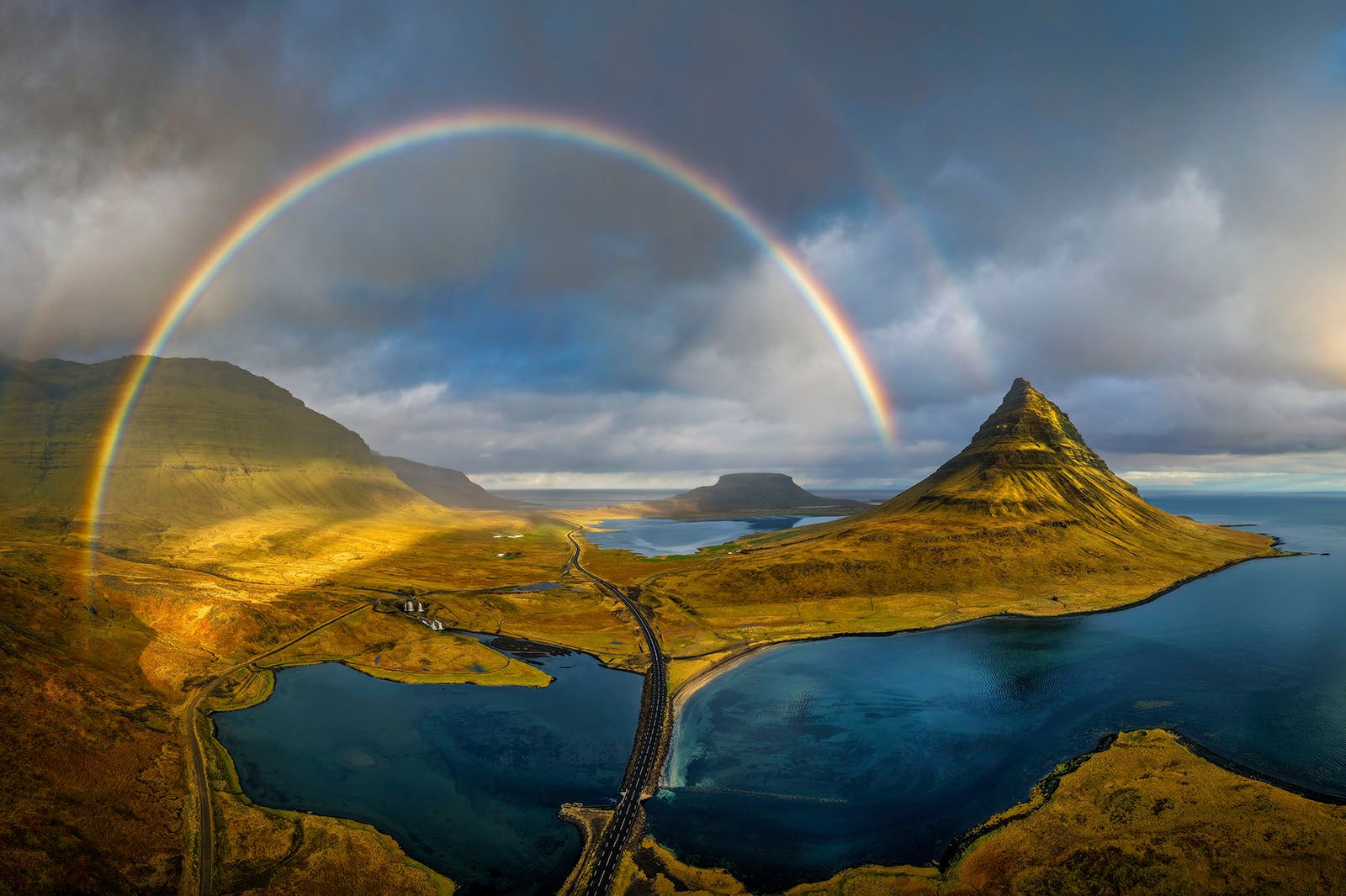 A vivid rainbow arches over a rugged, mossy landscape with a conical mountain near the coast, surrounded by winding roads, blue water, and dramatic clouds in Iceland.