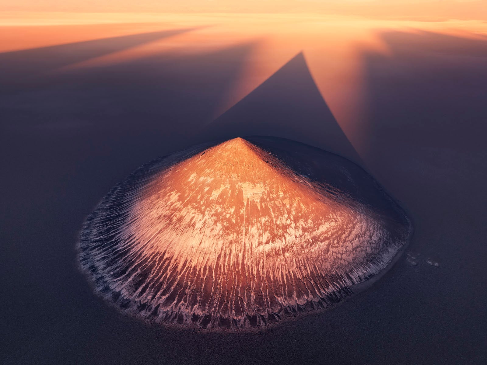 Aerial view of a solitary volcano with its peak glowing in warm sunlight, casting a long triangular shadow across dark, barren terrain at sunrise or sunset.