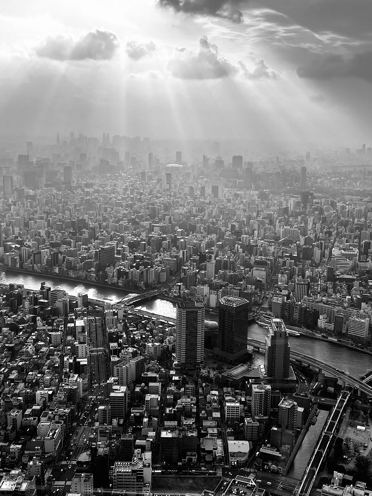 Aerial view of a dense cityscape with high-rise buildings and a river running through; rays of sunlight break through dramatic clouds, illuminating parts of the city. The image is in black and white.