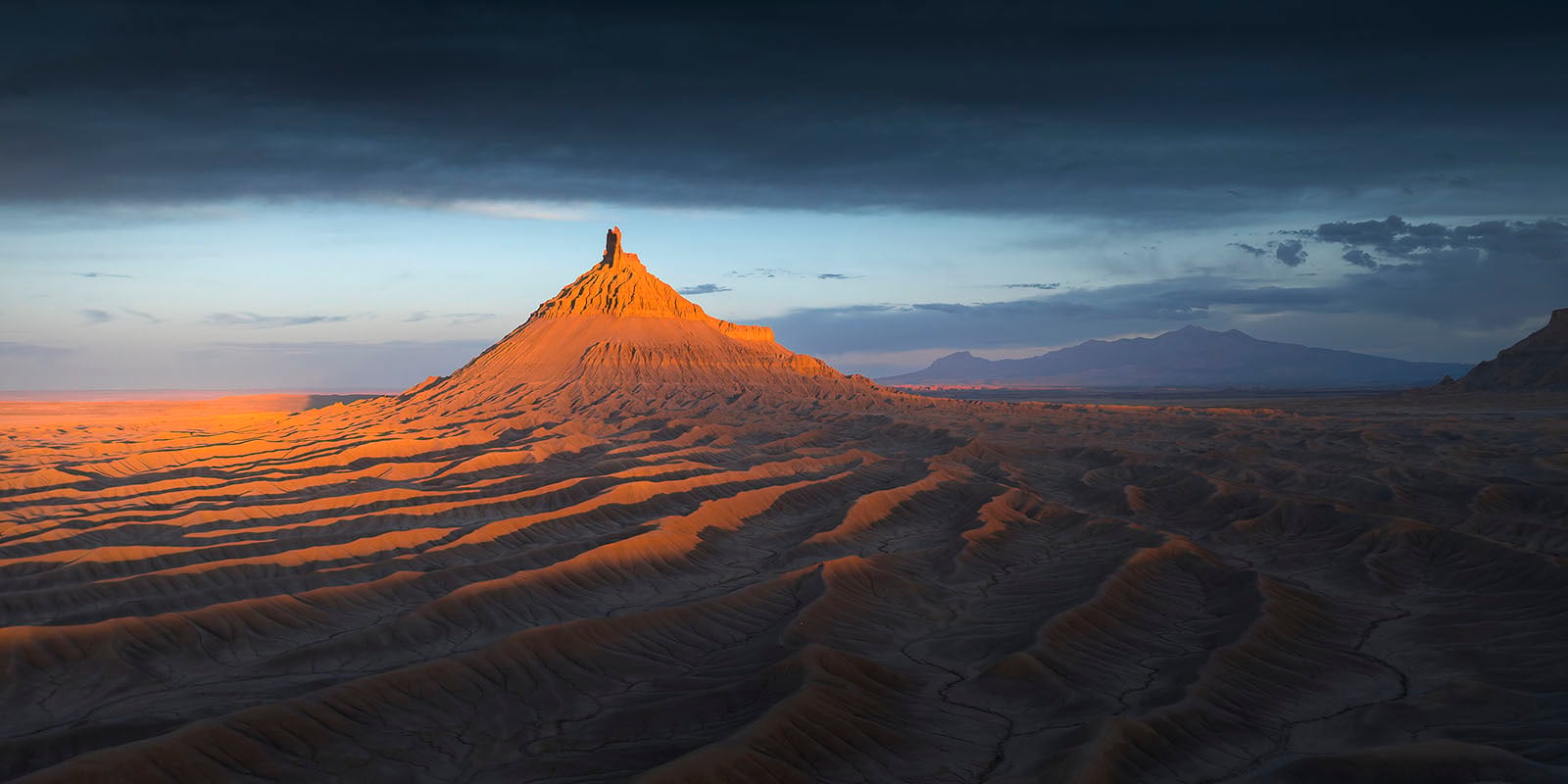 A dramatic desert landscape at sunset, featuring a pointed butte illuminated by golden sunlight, with ridged, barren terrain in the foreground and distant mountains under a cloudy sky.