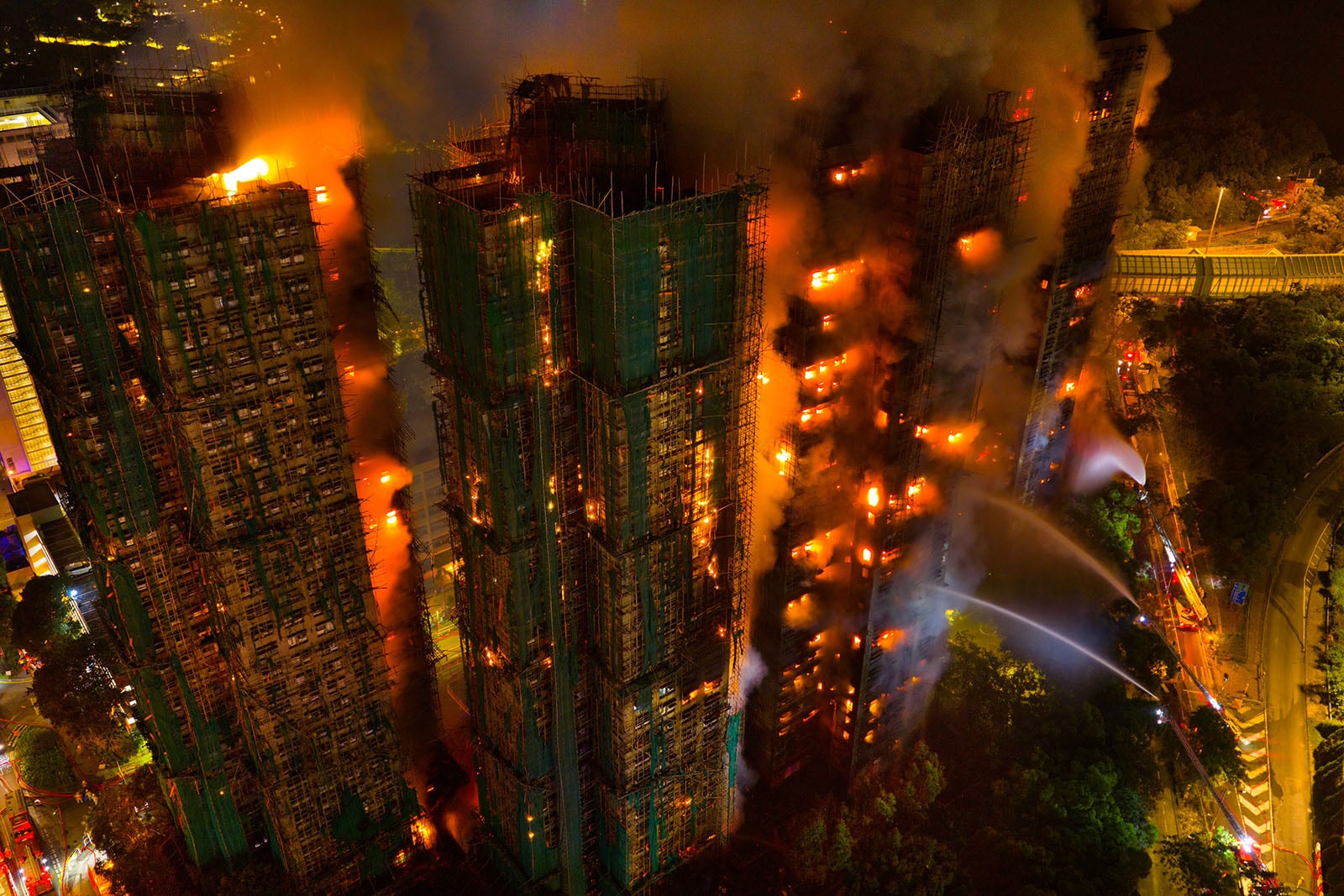 Aerial view of multiple high-rise buildings engulfed in flames at night, with firefighters spraying water and thick smoke billowing into the sky. Emergency vehicles and lights are visible on the ground below.