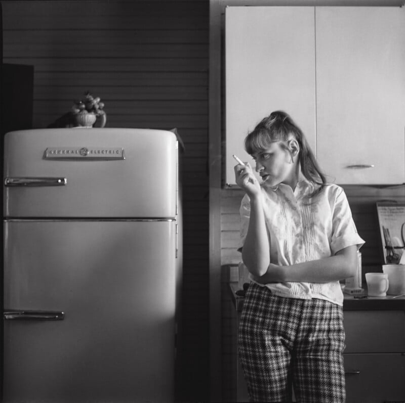 A young woman in plaid pants stands by a vintage refrigerator, holding a cigarette, with kitchen cabinets, a cup, and a small stuffed animal on top of the fridge visible in the background.