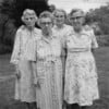 Four elderly women stand close together outdoors, wearing patterned dresses and glasses. They pose for a black-and-white photo, with trees and grass visible in the background.