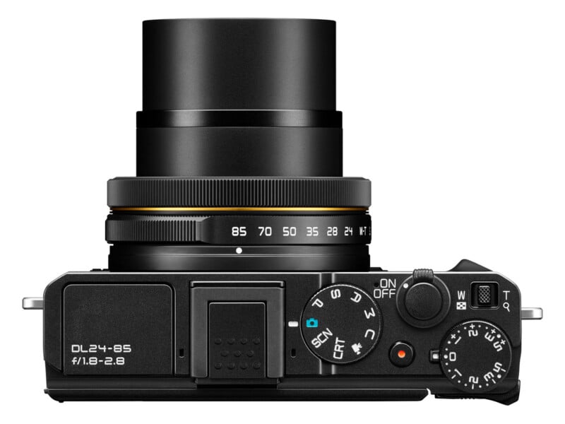Top view of a black digital camera with a protruding lens. The controls, dials, and labels such as "DL24-85" and "ON/OFF" are clearly visible, with the lens zoomed out and various settings marked.