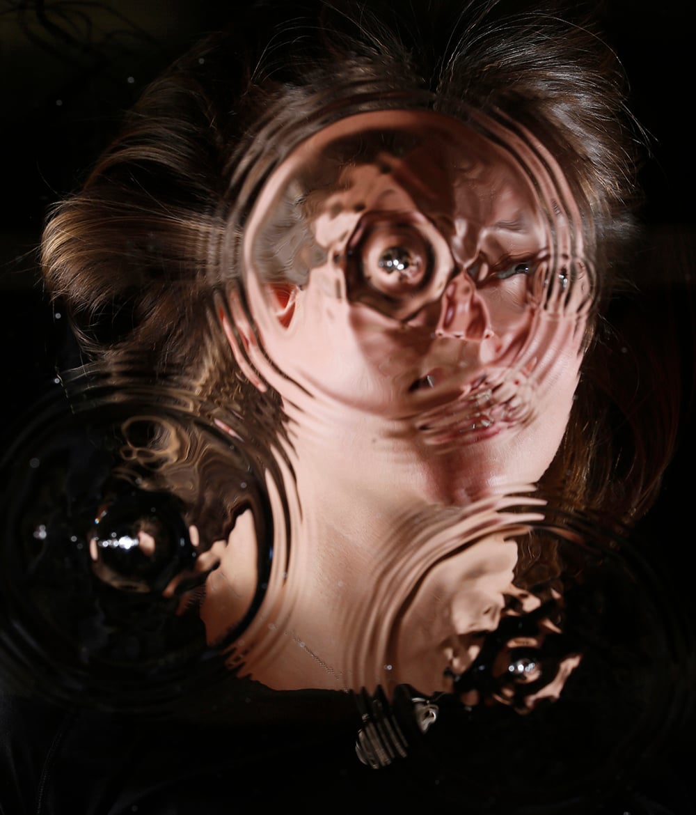 A woman's face is seen through glass with water droplets, creating a distorted and wavy reflection of her features and hair.