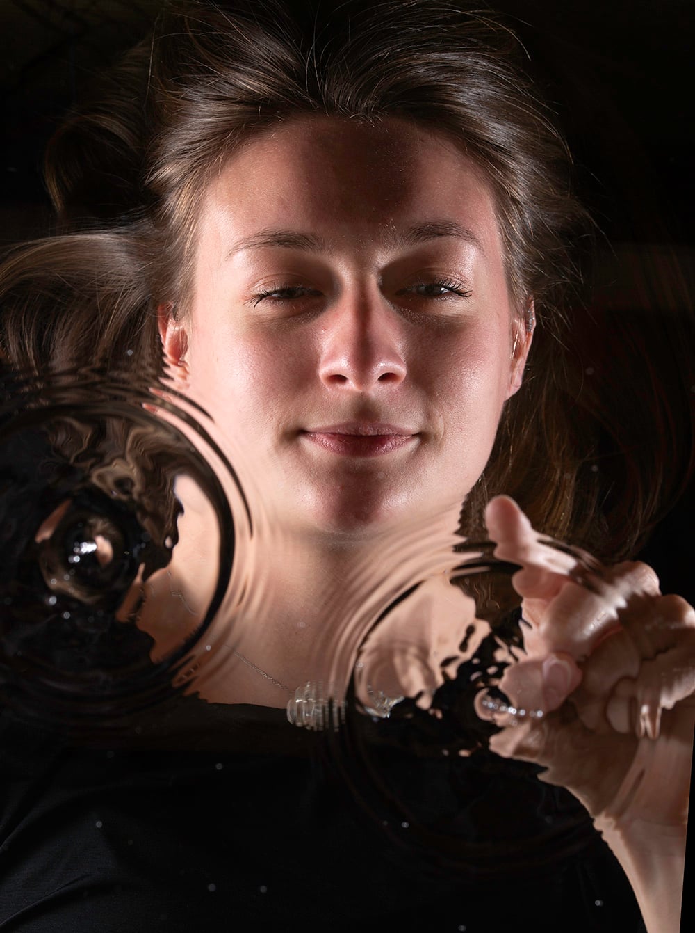 A woman with brown hair looks down and touches a transparent surface, creating circular ripples with her fingers, while light highlights her face and features.