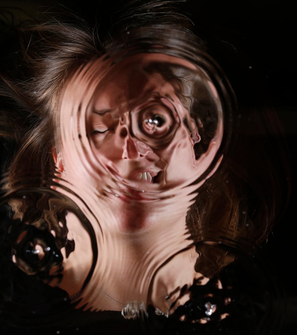 A woman's face is distorted by pressed glass or plastic, with ripples creating a warped, abstract effect. Her hair is spread out and she appears to be smiling behind the textured surface.