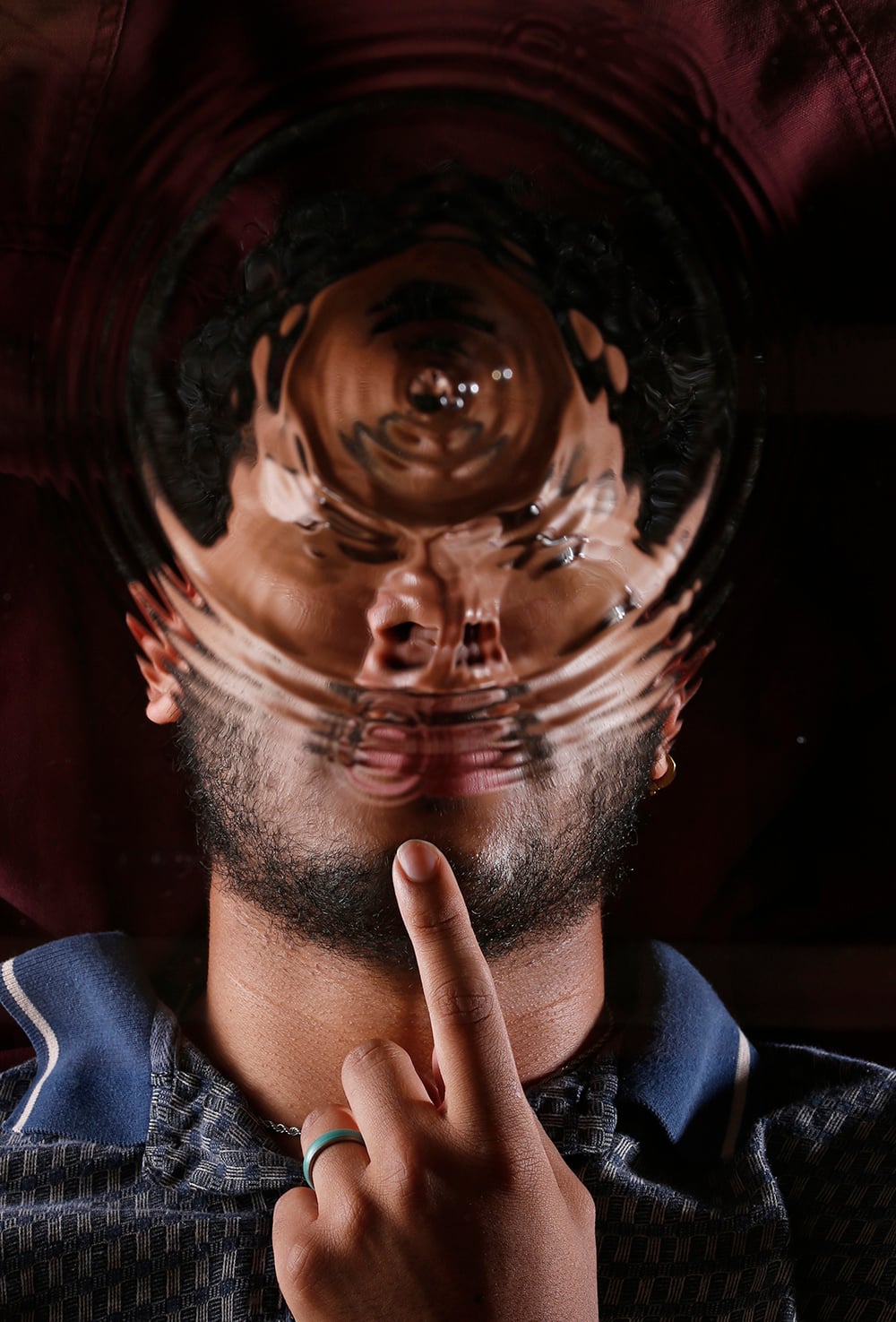 A person lies down touching their lips, with their face distorted by circular water ripples, creating a surreal, abstract effect.