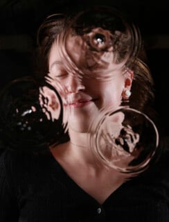 A woman stands behind rippled glass, her face partially distorted by three circular patterns, eyes closed and smiling softly, wearing a black top and pearl earrings against a dark background.