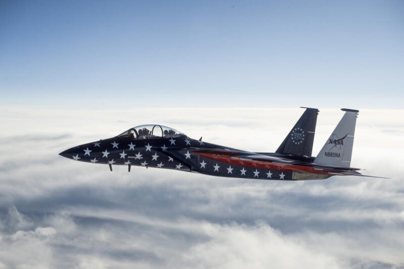 A fighter jet painted with stars and stripes, resembling the American flag, flies above the clouds in a clear sky.