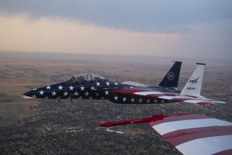 A fighter jet with a stars-and-stripes American flag design flies over a city at sunset, with another aircraft's wingtip visible in the foreground.